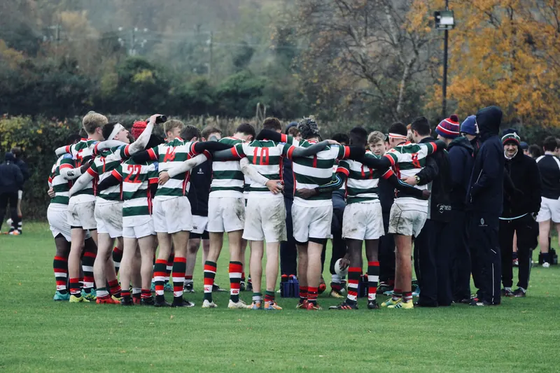 A rugby team in green, white and red hooped shirts stands in a huddle on a grass pitch with autumn trees behind.