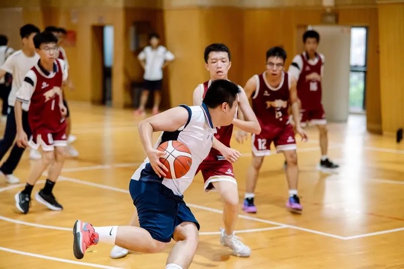 A teenage basketball player in white dribbles past defenders in maroon on an indoor wooden court.