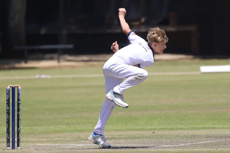 A young cricketer in whites leaps through his bowling action, with the stumps behind him on a sunny ground.