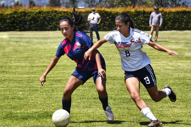 Two young female footballers, one in navy and pink and one in white, challenge for the ball on a sunny grass pitch.