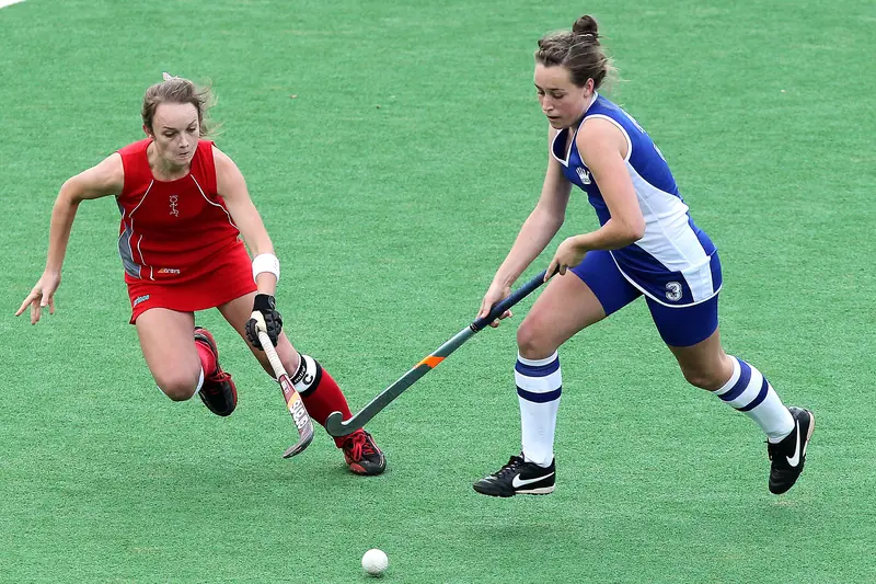 Two women playing hockey, one in red and one in blue and white, go for the ball on a green artificial pitch.