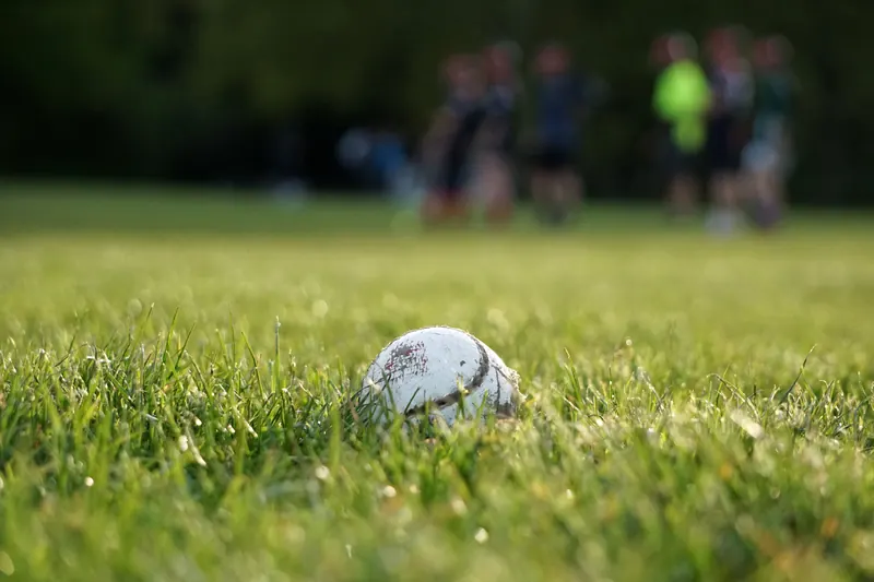 A worn hurling ball lies in the grass while players train out of focus in the background.