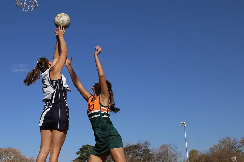 A netball player in navy and white shoots at the ring as a defender in green and orange jumps to block under a clear blue sky.