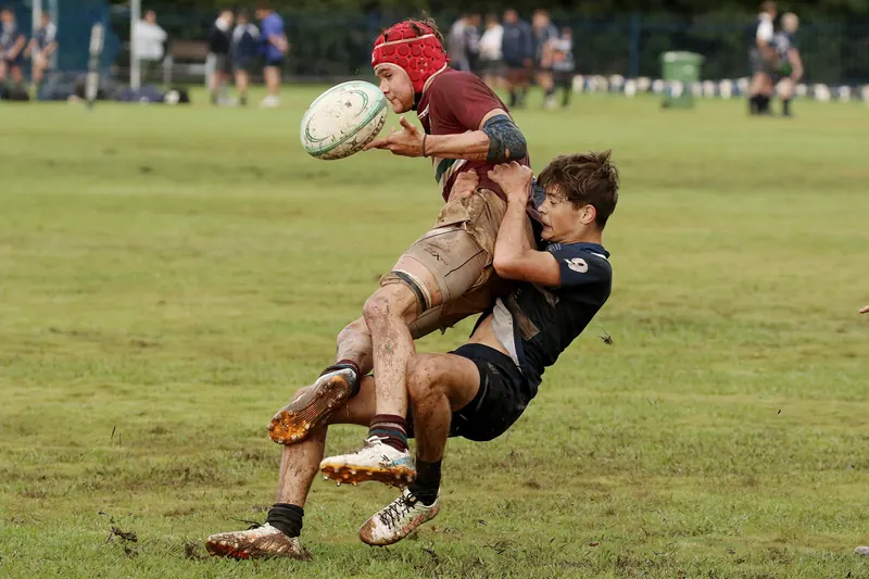 A young rugby player in a maroon shirt and red scrum cap is tackled as he passes the ball on a muddy pitch.