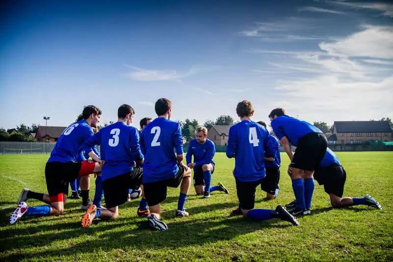 Adult footballers in blue kit kneel together on a sunny grass pitch before a match, with houses behind.