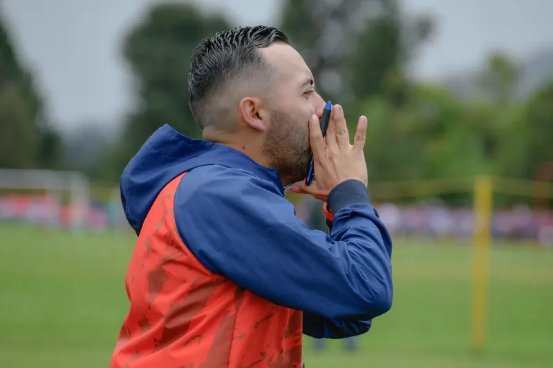A man in a blue and red jacket cups his hands to shout instructions from the side of a grass pitch.
