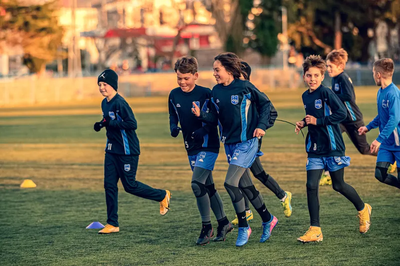 Children in navy and blue training kit jog together across a grass pitch in warm, low sunlight.